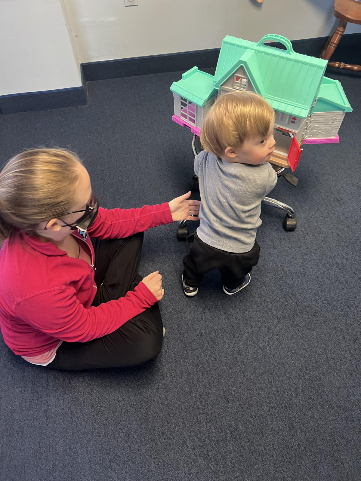 a small boy stands holding on to a doll house while a therapist prompts him
